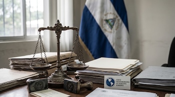 Primer plano de una balanza de bronce sobre una mesa de madera con documentos judiciales. A su lado, sellos, una tarjeta de la Corte IDH y la bandera de Nicaragua desenfocada.