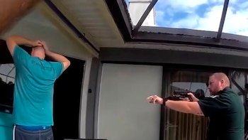 Un hombre de espaldas con camiseta azul verdoso y manos en la cabeza frente a un oficial de policía que apunta con un arma de fuego. Cielo nublado visible