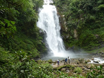 Una cascada de gran caudal desciende sobre rocas, rodeada de abundante vegetación. Tres personas caminan sobre piedras en el río que se forma en la base