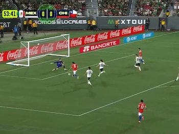 Jugadores de fútbol disputan un balón cerca del arco en un campo de juego, con un marcador en pantalla que indica México 0, Chile 0
