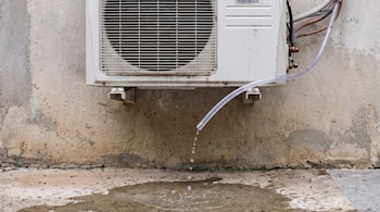 Unidad exterior de aire acondicionado con ventilador y rejilla, tuberías de cobre, un tubo de drenaje transparente goteando agua sobre un suelo de hormigón mojado junto a una pared.