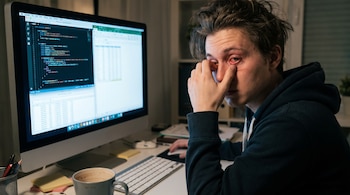 Hombre joven con ojos rojos e irritados, camiseta con capucha negra, sentado frente a un computador con código y hojas de cálculo en la pantalla, una taza de café.