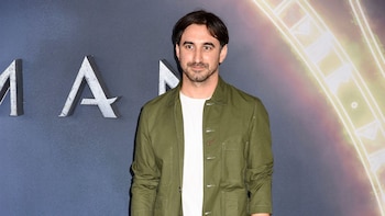 Un hombre con barba, vestido con blazer de tweed marrón, camiseta blanca, pantalón marrón y cinturón oscuro, posa frente a un fondo gris con logo de Apple TV+