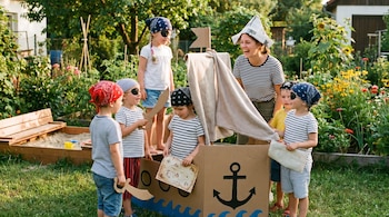 Un grupo de seis niños y una maestra sonríen jugando a los piratas con un barco de cartón. Los niños llevan bandanas y algunos parches de ojo en un jardín soleado.