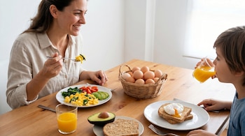 Una mujer y un niño desayunando en una mesa de madera. La mujer come huevos revueltos con espinacas. El niño bebe zumo de naranja. Hay tostadas, aguacate y una cesta de huevos.