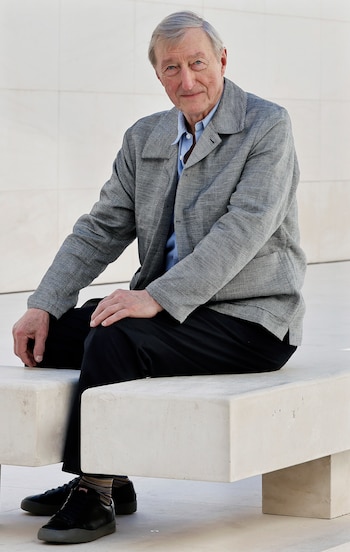 Julian Barnes sentado en un banco de hormigón, vestido con una camisa azul claro, chaqueta gris y pantalones negros, mirando a cámara