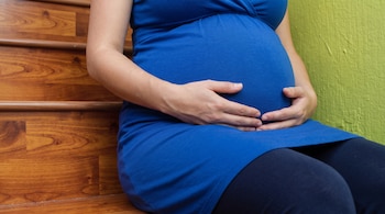 Mujer embarazada vestida de azul sentada en escaleras de madera. Manos de la mujer sobre el vientre abultado. Pared verde al fondo.