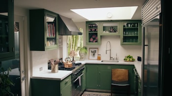 Cocina luminosa con gabinetes verdes, encimeras de mármol blanco, estufa de acero inoxidable y fregadero. Se ven utensilios, limas, y una tetera