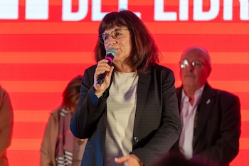 Una mujer con cabello oscuro y anteojos sostiene un micrófono, viste saco negro y blusa blanca, con personas borrosas y fondo rojo con letras blancas