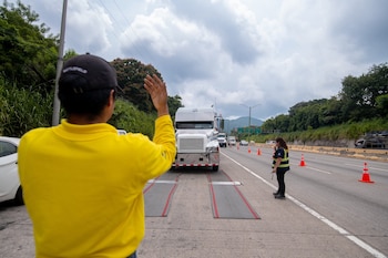 Un hombre con camisa amarilla hace señas a un camión blanco colocado sobre plataformas de pesaje en una carretera con conos de tráfico