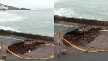 Forado grande en una carretera costera asfaltada, con rocas expuestas, cercado con malla de seguridad naranja y cinta amarilla. El mar agitado se ve al fondo