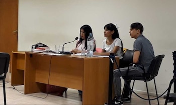 Una mujer habla por un micrófono en una mesa de madera con vasos y libros. A su lado, otra mujer y un hombre escuchan, sentados en sillas