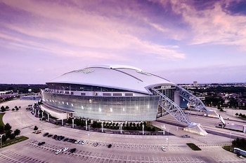 Vista aérea de un estadio moderno con techo blanco y fachada de vidrio, rodeado de un amplio estacionamiento bajo un cielo púrpura y anaranjado