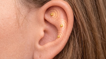 Primer plano de una oreja humana con tres pequeñas esferas doradas adheridas y anillos concéntricos luminosos a su alrededor. Se observa cabello castaño.