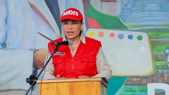 Una mujer con gorra y chaleco rojos habla ante un micrófono sobre un atril de madera con un mural colorido al fondo