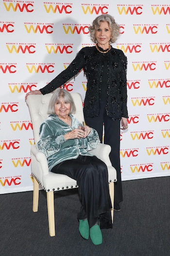 Robin Morgan posa sentada en un sillón junto a Jane Fonda, de pie tras ella, ante un telón con logotipos de Women's Media Center