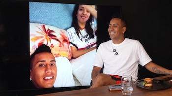 Christian Cueva sentado frente a una mesa con un vaso de agua, una hamburguesa y un teléfono, con una pantalla que muestra a Christian Cueva y Rosángela Espinoza.