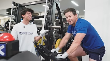 Mark Wahlberg en un gimnasio, sonriendo mientras pedalea en una máquina de ejercicio, con guantes blancos. Un hombre joven de camiseta blanca sonríe a su lado