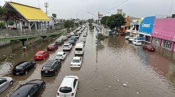 Vista aérea de una avenida urbana inundada por agua marrón, con una larga fila de vehículos detenidos y edificaciones a los lados.