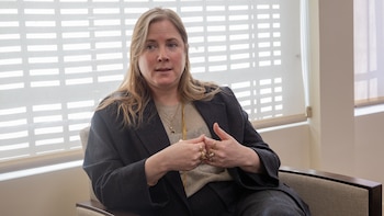 Retrato de primer plano de Dana Erlich, una mujer de cabello rubio, ojos azules y piel clara, usando chaqueta oscura y joyas doradas, con fondo blanco y rejilla