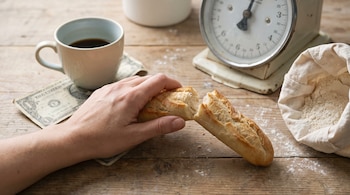 Mano sujetando pan francés partido sobre mesa de madera. Hay una taza de café, un billete de un dólar, una balanza de cocina y una bolsa de harina.