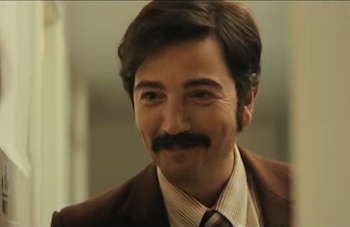 Primer plano de un hombre caucásico de mediana edad con cabello oscuro y bigote, vistiendo traje marrón y camisa a rayas, sonriendo
