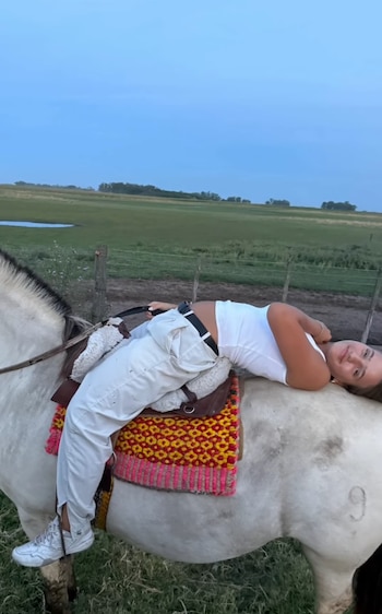 Joven mujer con top blanco y pantalones claros recostada sobre el lomo de un caballo blanco con silla de montar y manta de colores, en un campo verde