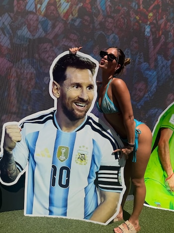 Una mujer con bikini celeste y gafas de sol posa haciendo un gesto de beso al lado de una figura de cartón de Lionel Messi