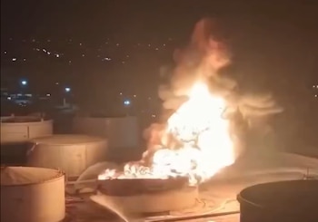 Fuego ardiendo sobre un tanque industrial cilíndrico. Varios tanques similares alrededor. Chorro de agua intentando apagarlo. Cielo nocturno con luces de ciudad a lo lejos