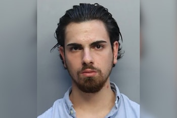 Retrato de un hombre joven con cabello oscuro, barba y una camisa azul claro frente a un fondo gris liso. Tiene una marca roja en el cuello