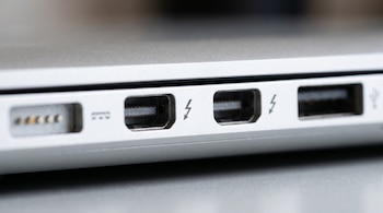 Primer plano del lateral de un ordenador portátil plateado que muestra un puerto de carga MagSafe, dos puertos Thunderbolt y un puerto USB-A.