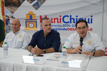 Tres hombres sentados en mesa blanca con carteles de nombres y botellas de agua. Uno sujeta micrófono. Fondo con banner y un logo de edificio