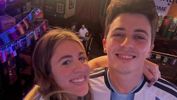 Primer plano de un hombre y una mujer sonriendo, el hombre con camiseta blanca y celeste de fútbol, la mujer abrazándolo, en el fondo un bar con banderas