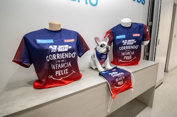 Dos camisetas deportivas azul y rojo sobre maniquíes, una figura de perro con pañuelo y una bolsa con cordón sobre una mesa blanca