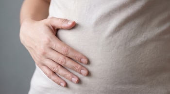Primer plano de una persona, con una mano apoyada sobre su abdomen cubierto por una camiseta de color claro, con un fondo gris liso.