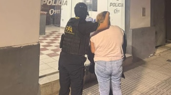 Oficial de la FPA con chaleco oscuro sujeta a una mujer de cabello claro con camiseta rosa y jeans. Al fondo, la entrada de una comisaría con el logo 'POLICIA 0911'