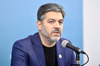Primer plano de Carlos Bianco, un hombre con cabello gris y barba, vestido con una chaqueta azul y suéter negro, hablando en un micrófono