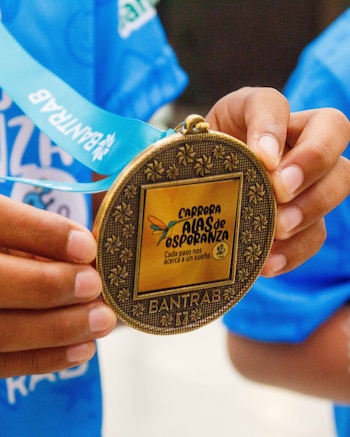 Manos sujetan una medalla redonda color bronce con texto "Carrera Alas de Esperanza", un ave estilizada y el nombre Bantrab en la medalla y cinta azul