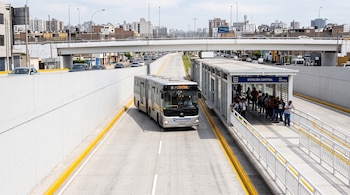 Autobús articulado gris en carril exclusivo de concreto junto a una estación de pasajeros y un puente al fondo.