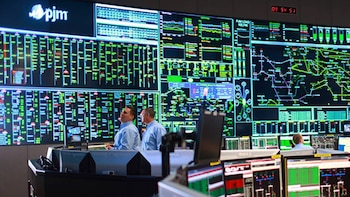 Sala de control con pantallas gigantes mostrando gráficos y mapas de red eléctrica. Dos hombres de camisa azul observan. Mesas con múltiples monitores de computadora