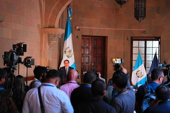 Un hombre vestido de traje habla en un podio frente a un micrófono. Detrás, la bandera de Guatemala y otra oscura. Cámaras y público asisten
