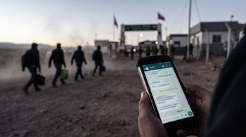 Manos sostienen un teléfono celular con una aplicación de mensajería frente a varias personas caminando en un paso fronterizo.