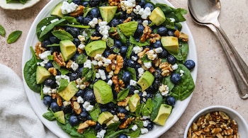 Plato blanco lleno de una ensalada fresca con espinacas, arándanos, trozos de aguacate, nueces, queso feta y hojas de menta. Cucharas y un cuenco de nueces al lado.