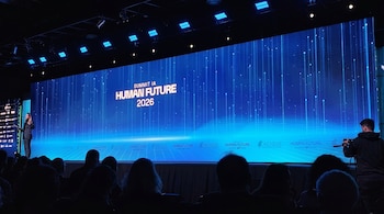 Vista de un escenario con una gran pantalla azul que muestra 'SUMMIT IA HUMAN FUTURE 2026'. Una oradora está a la izquierda y un público sentado en primer plano.