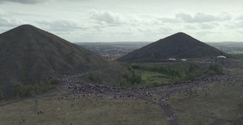 Dos colinas oscuras y cónicas rodeadas de caminos con numerosas personas en una zona llana bajo un cielo con nubes