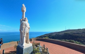 Estatua de explorador con armadura sobre una base oscura, con el océano y el cielo azul al fondo. A la derecha, una colina verde. Una persona a la izquierda
