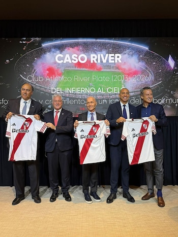 Cinco hombres con traje posan de pie, sosteniendo camisetas blancas de fútbol con una franja roja. Detrás, una pantalla muestra un estadio lleno con humo rojo y el texto "CASO RIVER"