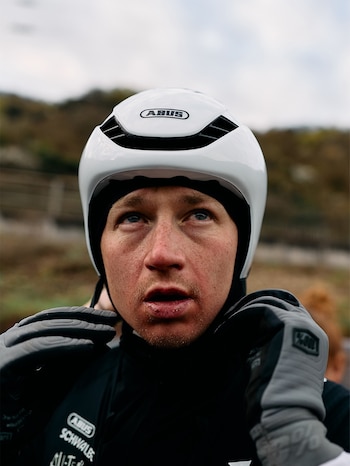 Hombre con casco blanco ABUS, chaqueta negra y guantes grises se ajusta el equipo, con el rostro serio y la mirada hacia arriba