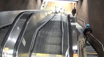 Una escalera mecánica y escaleras fijas paralelas en una estación con paredes de ladrillo; personas suben, una con sombrero azul y mochila