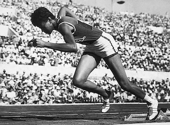 Fotografía en blanco y negro de Wilma Rudolph en una pista de atletismo, en posición de salida con tacos. Fondo con gradas llenas de gente